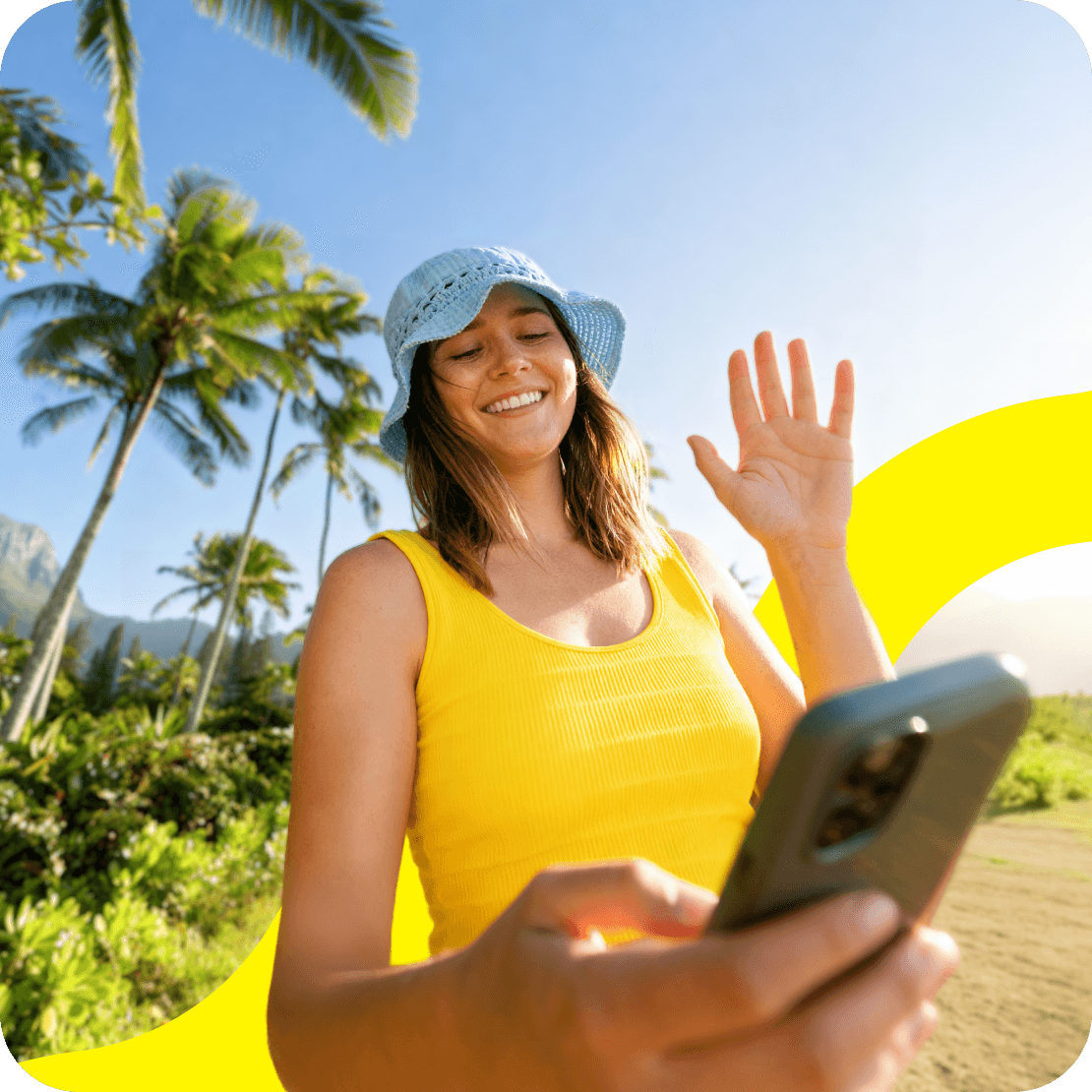 A young woman waving one hand and holding a phone in the other, with palm trees and mountains in the background.