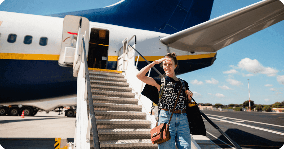 A woman with a backpack and a carry-on bag standing at the bottom of airplane stairs.