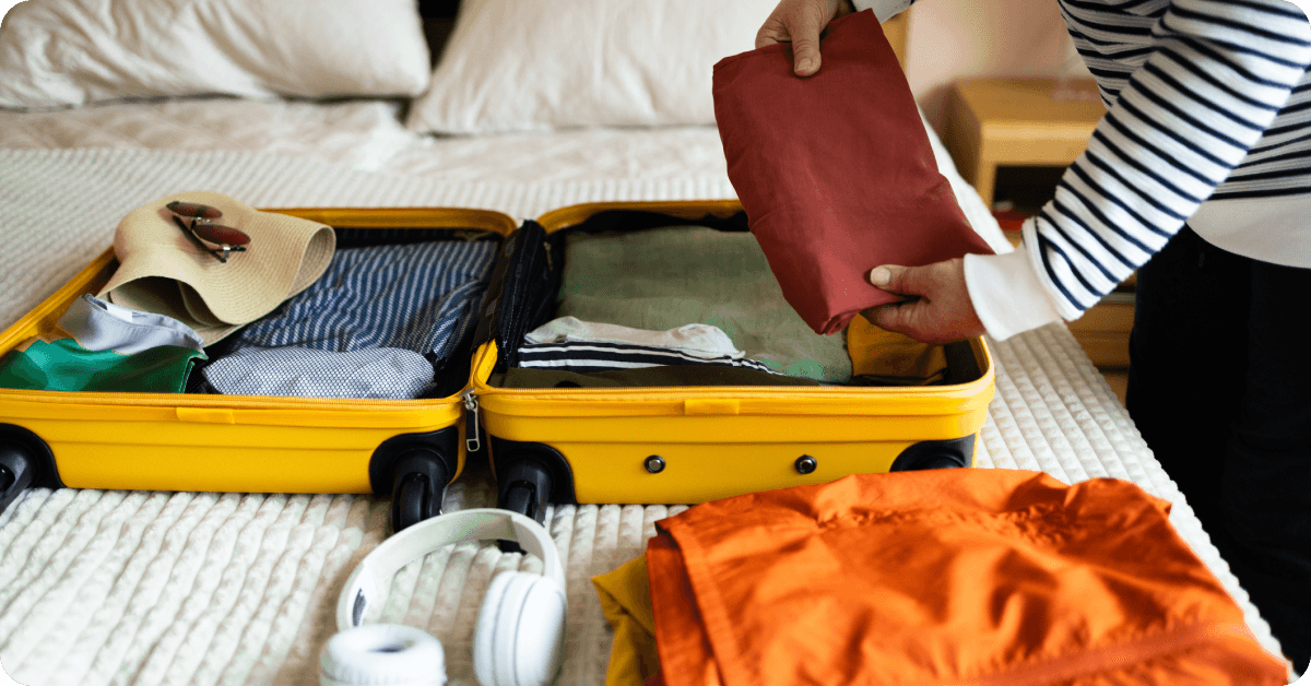 A person packing a yellow suitcase on a bed, with clothes and headphones laid out nearby.