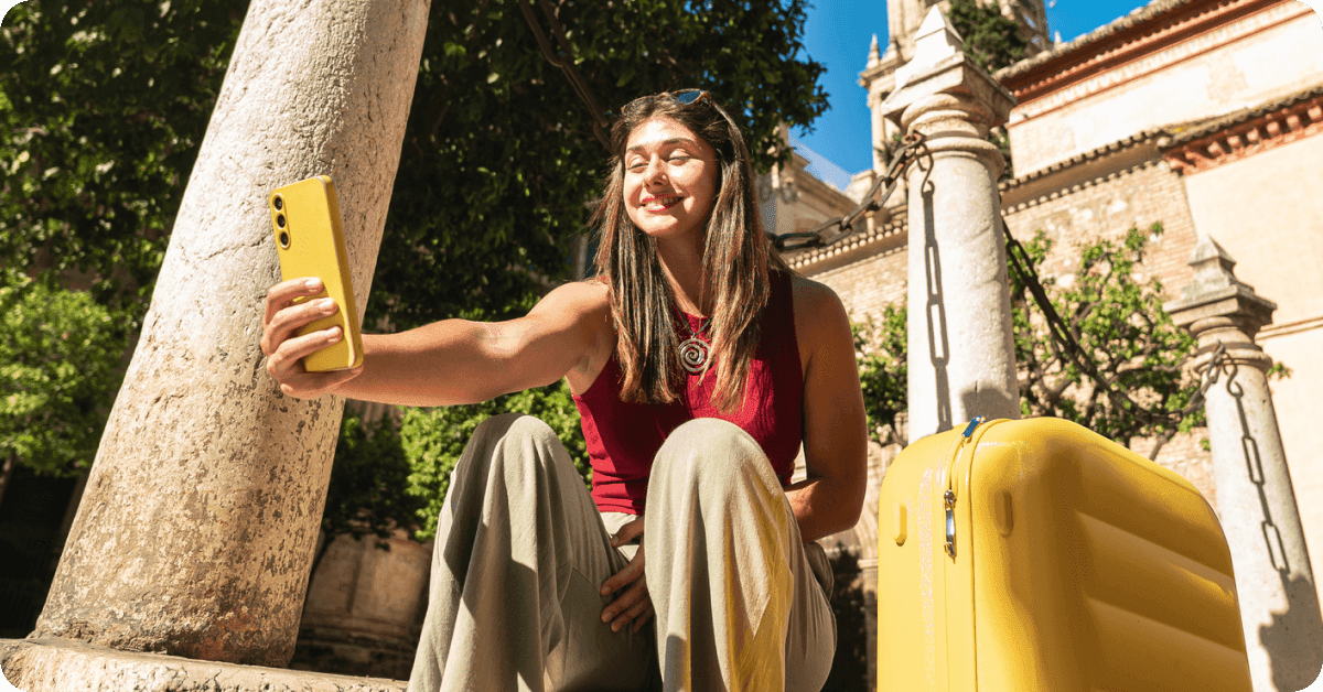 Young woman sitting on a pavement and taking a selfie, with a luggage nearby.