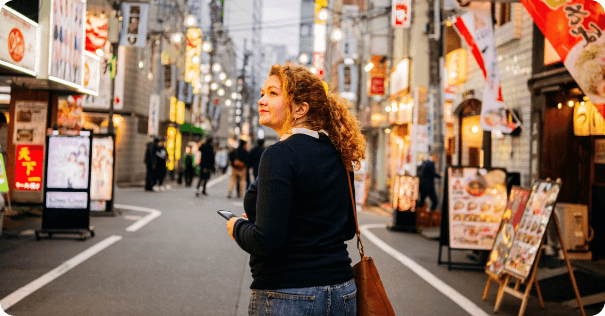 A person holding a phone while looking for a manga café entrance on a busy Japanese street.